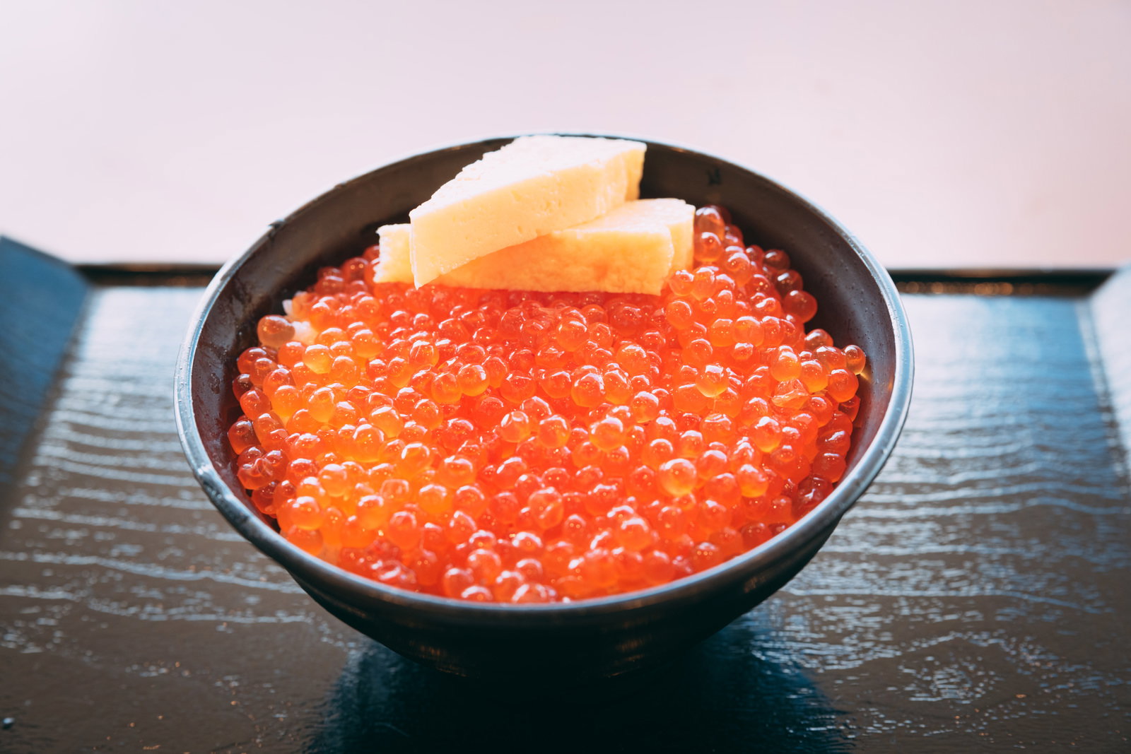 黒い丼に盛られた赤いイクラがぎっしり詰まったイクラ丼と、玉ねぎ、漬けたくあんが添えられている