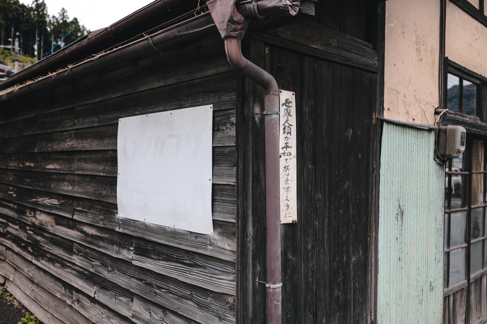 古い木造建物の壁に掲げられたマルフクの看板と平和を願う木製札