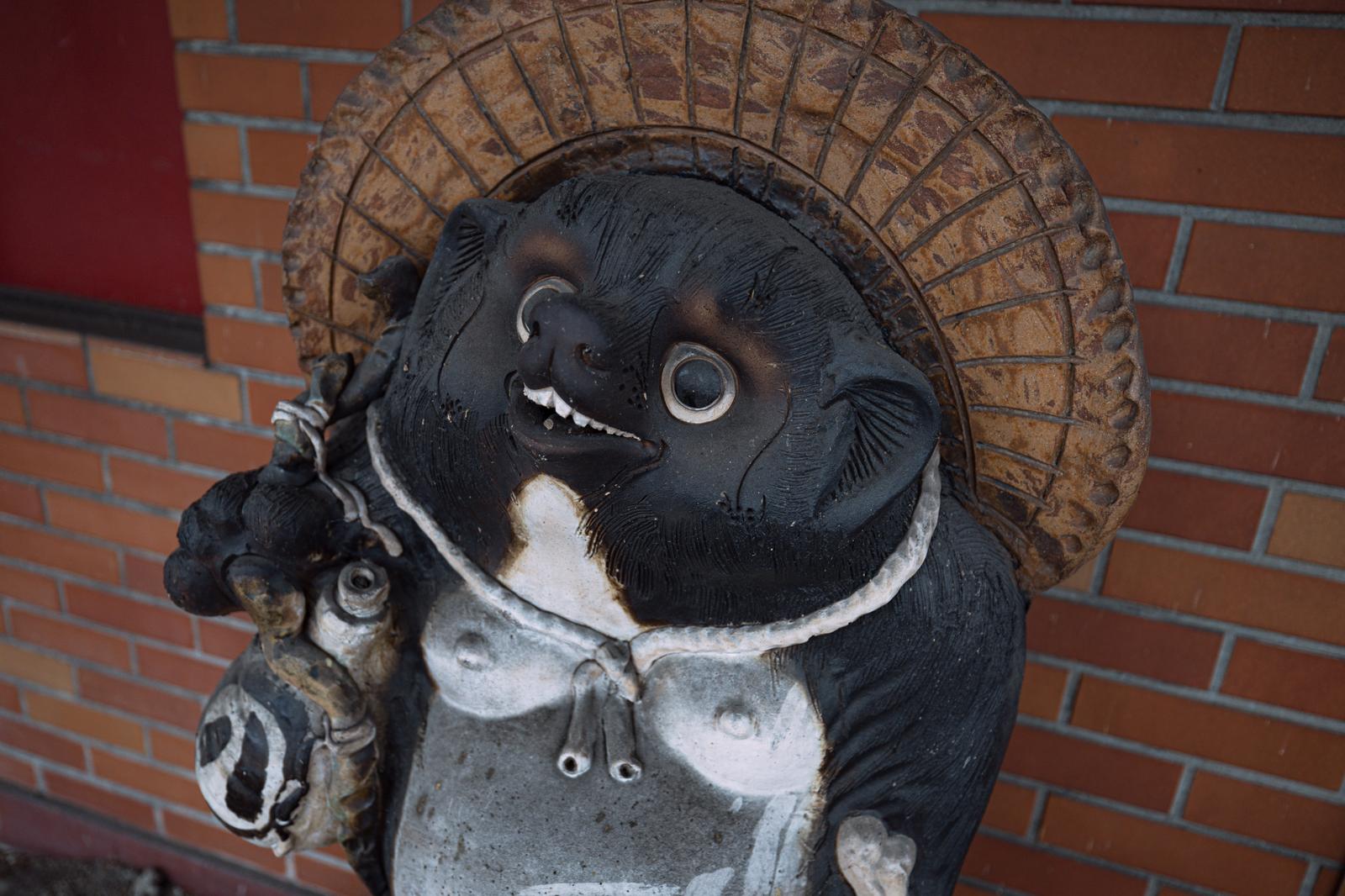 レンガ壁を背景に置かれた黒いたぬき置物。白い大きな目と笑顔が特徴