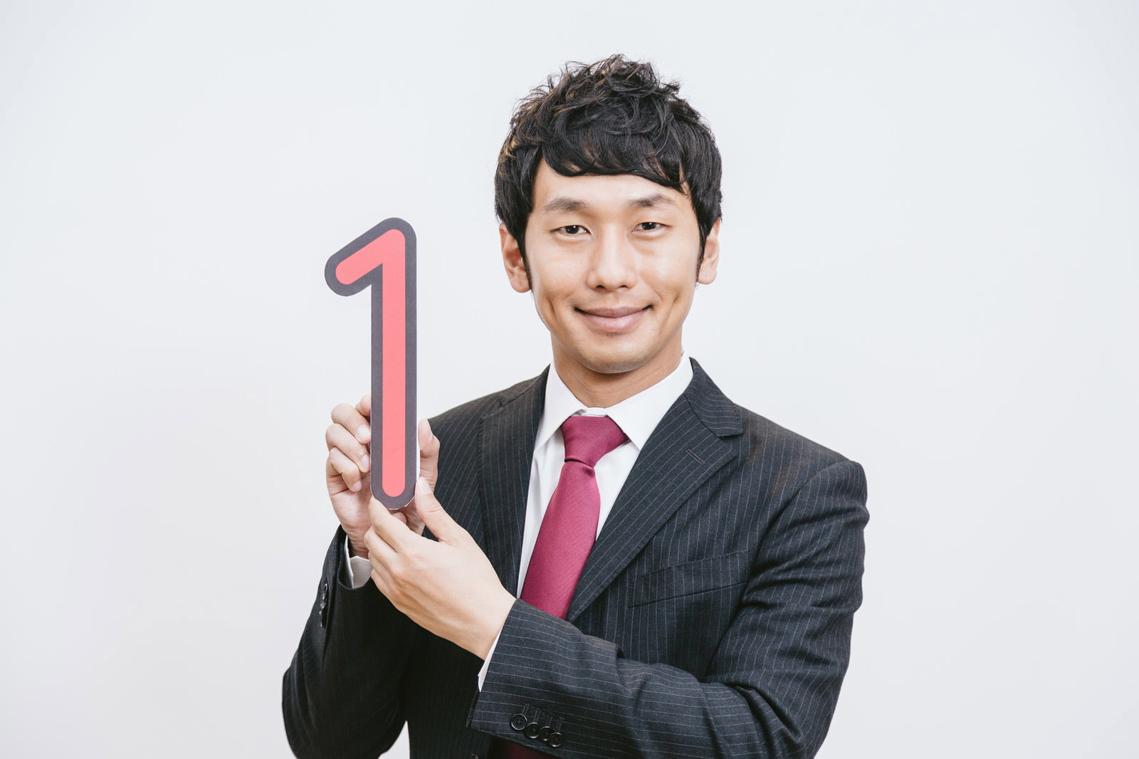 黒いストライプスーツとピンク色のネクタイをした男性がピンク色の数字1を両手で掲げて笑顔で正面を向いている［モデル：大川竜弥］