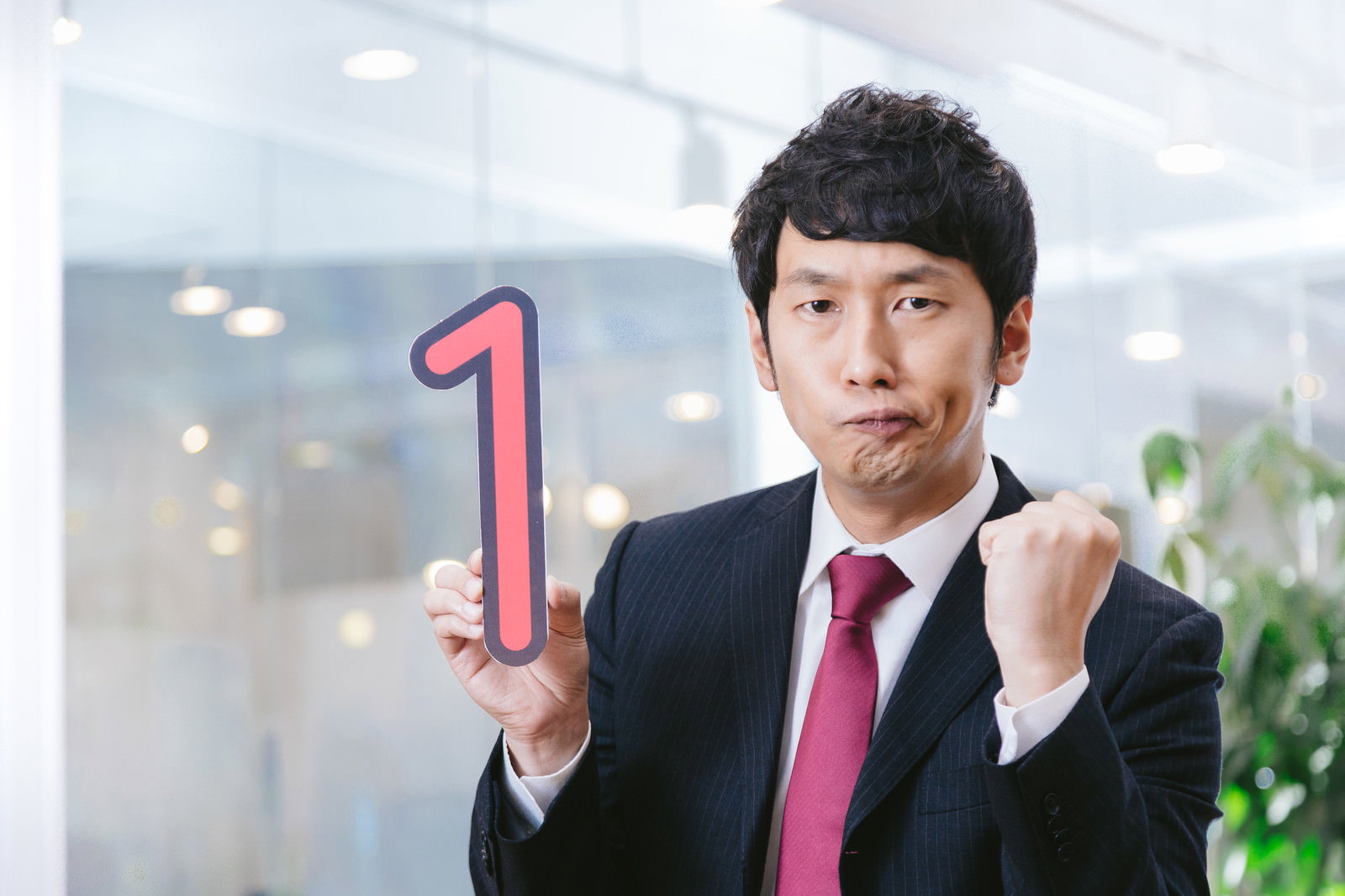 スーツ姿のビジネスマンが赤い「1」のカードを持ちながら拳を握るオフィスでのポーズ［モデル：大川竜弥］