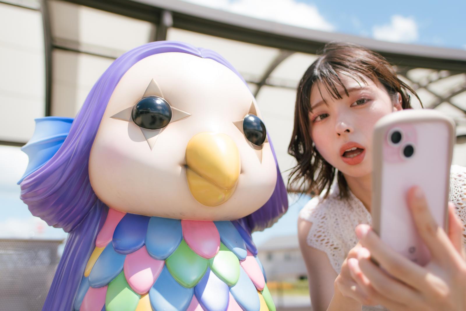 スマホを持つ女性の横で無関心な表情のアマビエの妖怪ベンチ［モデル：高尾実生］
