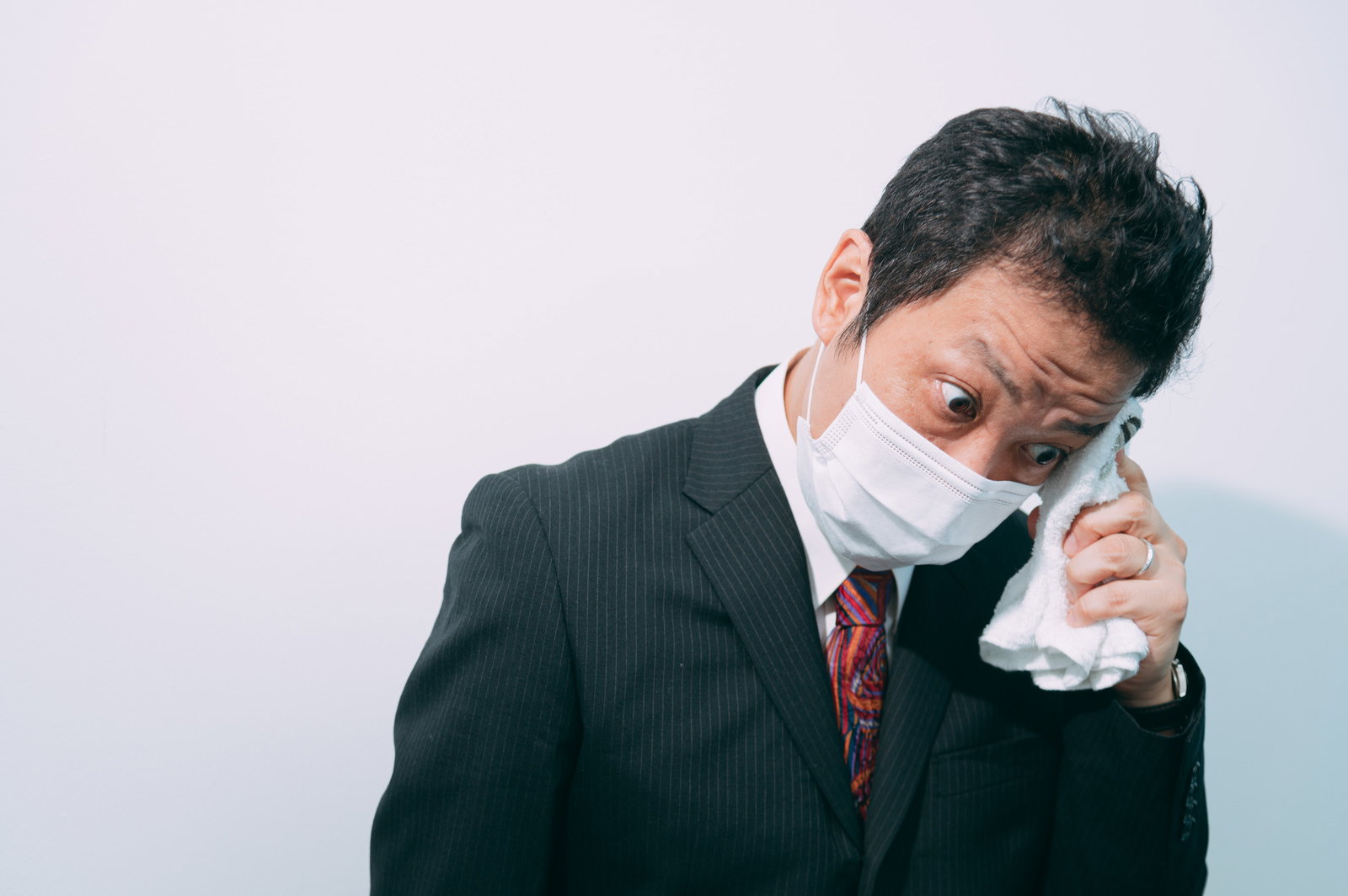 白いマスクを着けた黒いスーツの男性がハンカチで汗を拭いている［モデル：のせさん］