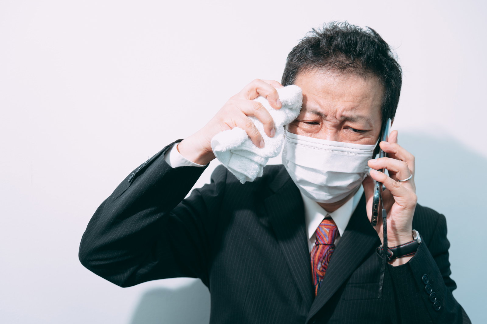 汗をハンカチで拭いながら電話で話すマスク着用の会社員男性［モデル：のせさん］