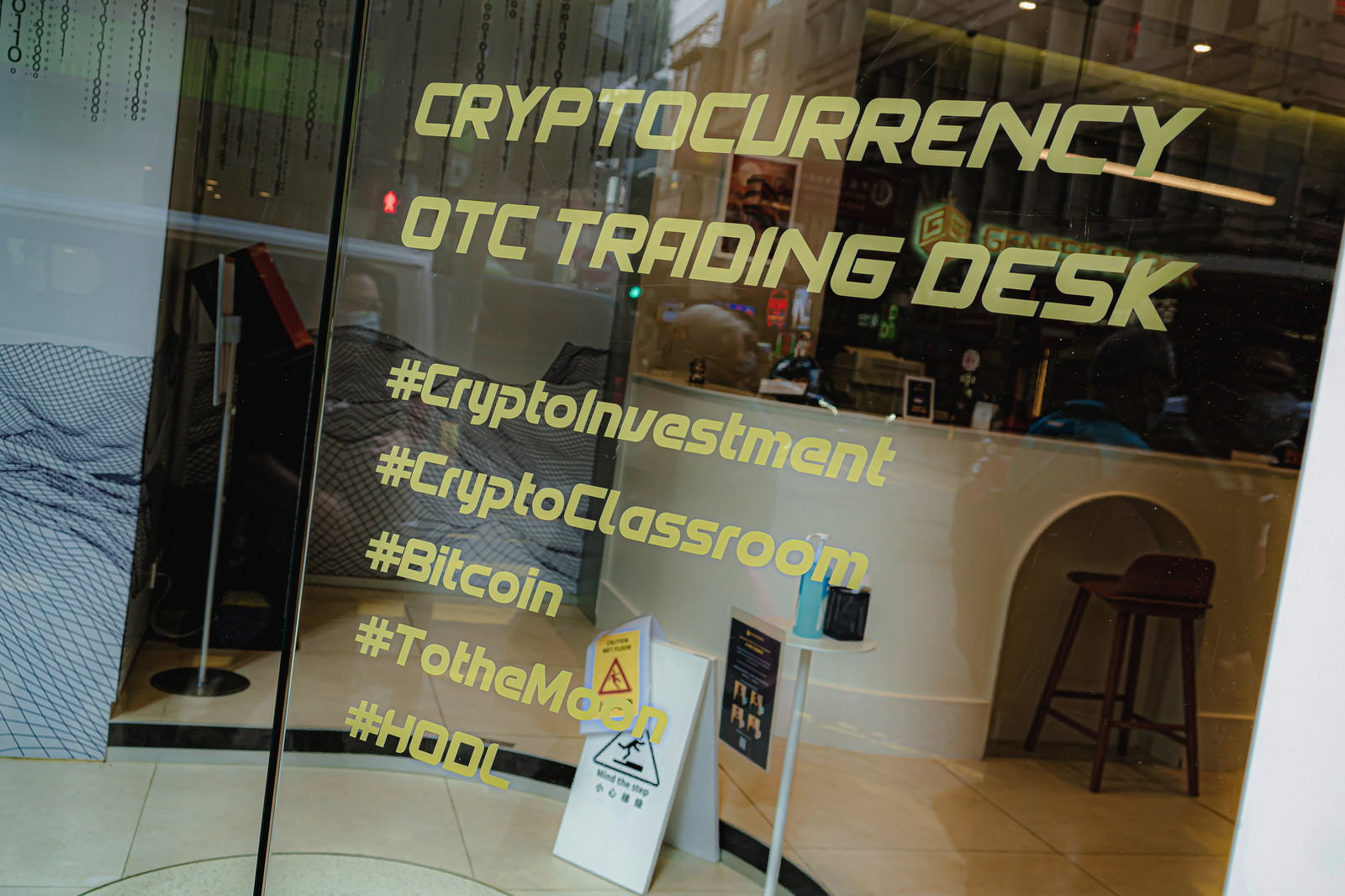 「CRYPTOCURRENCY OTC TRADING DESK」と表記された黄色い文字の店舗看板と仮想通貨関連のハッシュタグ