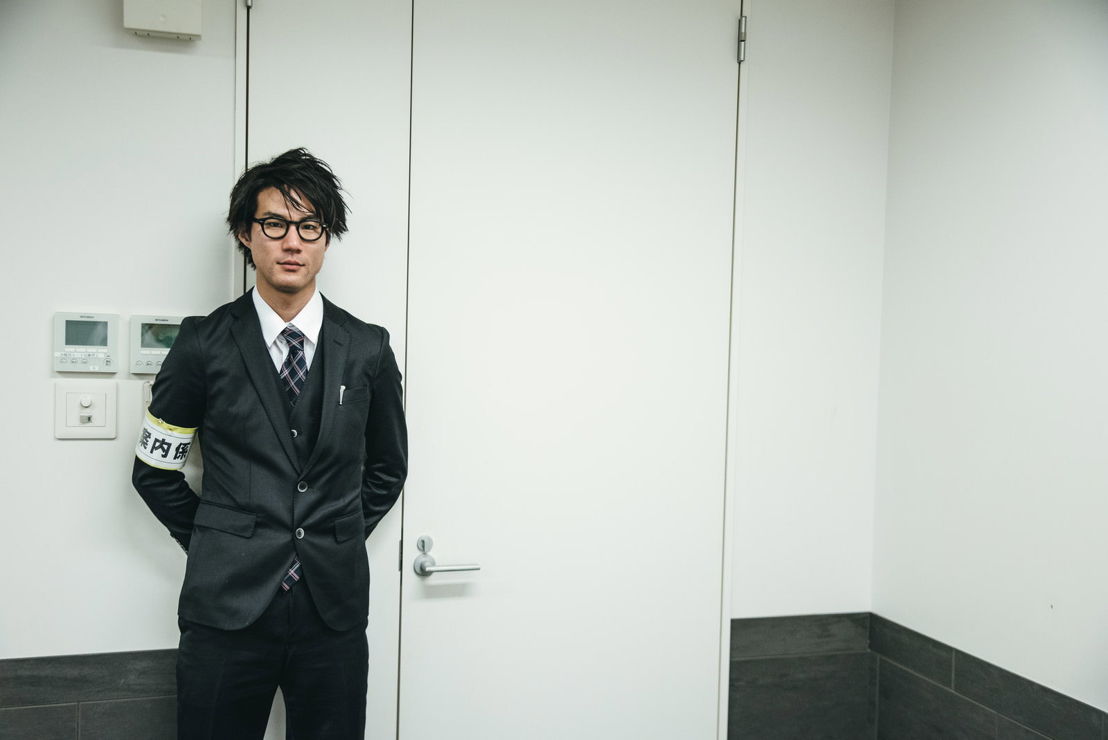 腕章をつけたスーツ姿の男性案内係が白い壁の前に立っている［モデル：村田裕章］