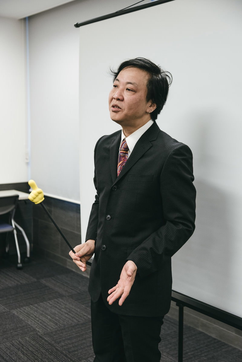 黒いスーツを着た男性講師が会議室で手を広げて講義している［モデル：のせさん］