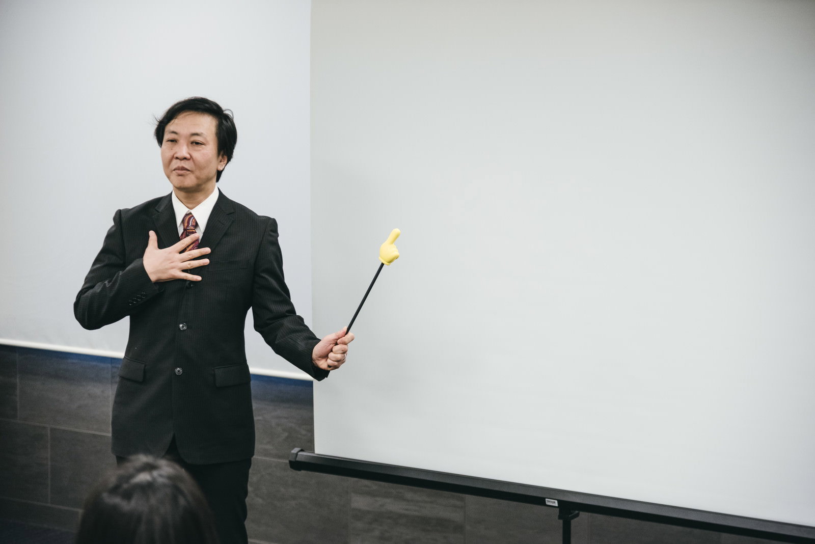黒いスーツを着た男性講師がホワイトボードを指し棒で指しながら説明している様子［モデル：のせさん］