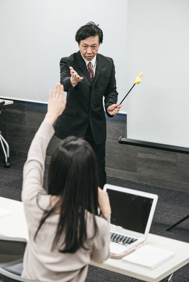 教室で受講者の挙手に応えて指名する黒いスーツ姿の男性講師［モデル：のせさん］