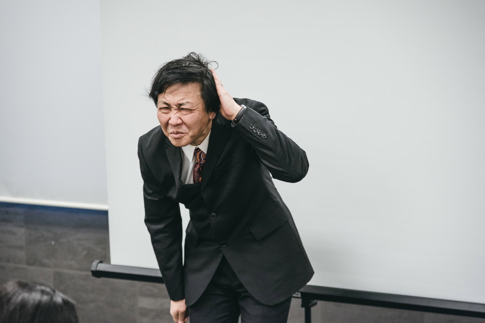 会議に遅刻して頭を下げて謝罪する黒いスーツ姿の部長［モデル：のせさん］