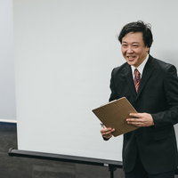 黒いスーツで資料を手に笑顔でセミナー参加者と対話する塾長の写真