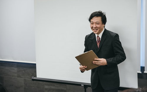 黒いスーツで資料を手に笑顔でセミナー参加者と対話する塾長