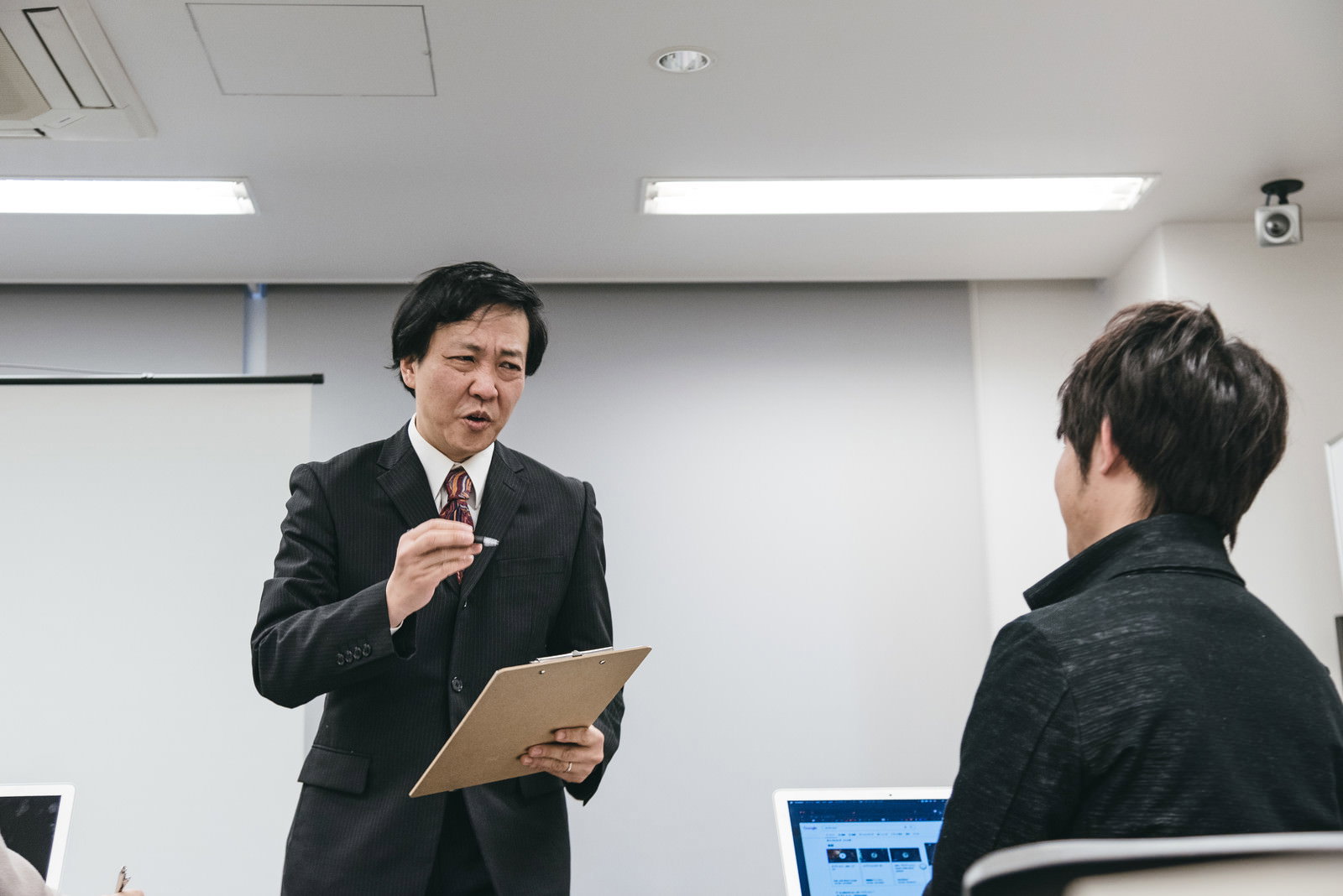 セミナー会場でスーツ姿の男性職員が資料を持って受講者に質問している様子［モデル：のせさん］