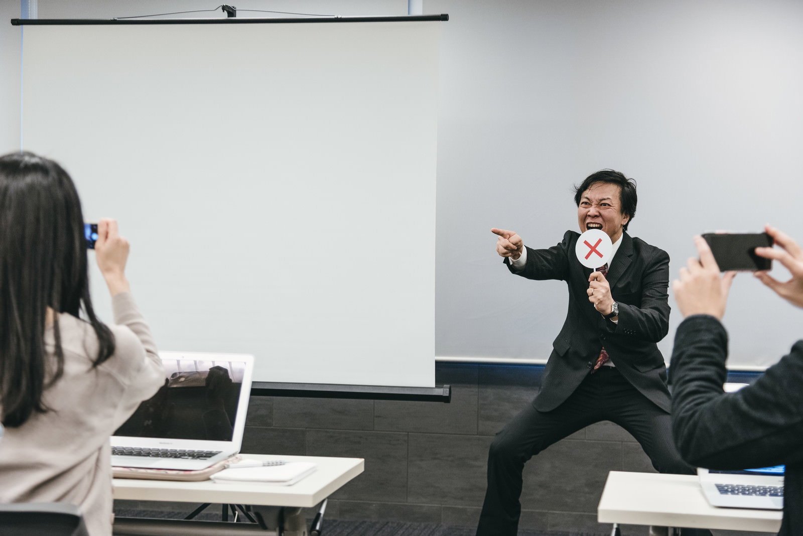 ホワイトボードの前で困惑した表情を見せるスーツ姿の男性講師［モデル：のせさん］