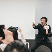 スーツ姿の講師がバツを出す様子をスマートフォンで撮影する参加者の写真