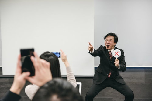 スーツ姿の講師がバツを出す様子をスマートフォンで撮影する参加者