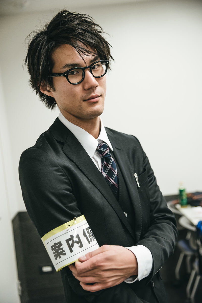 腕章をつけて案内業務を行うスーツ姿の男性職員［モデル：村田裕章］
