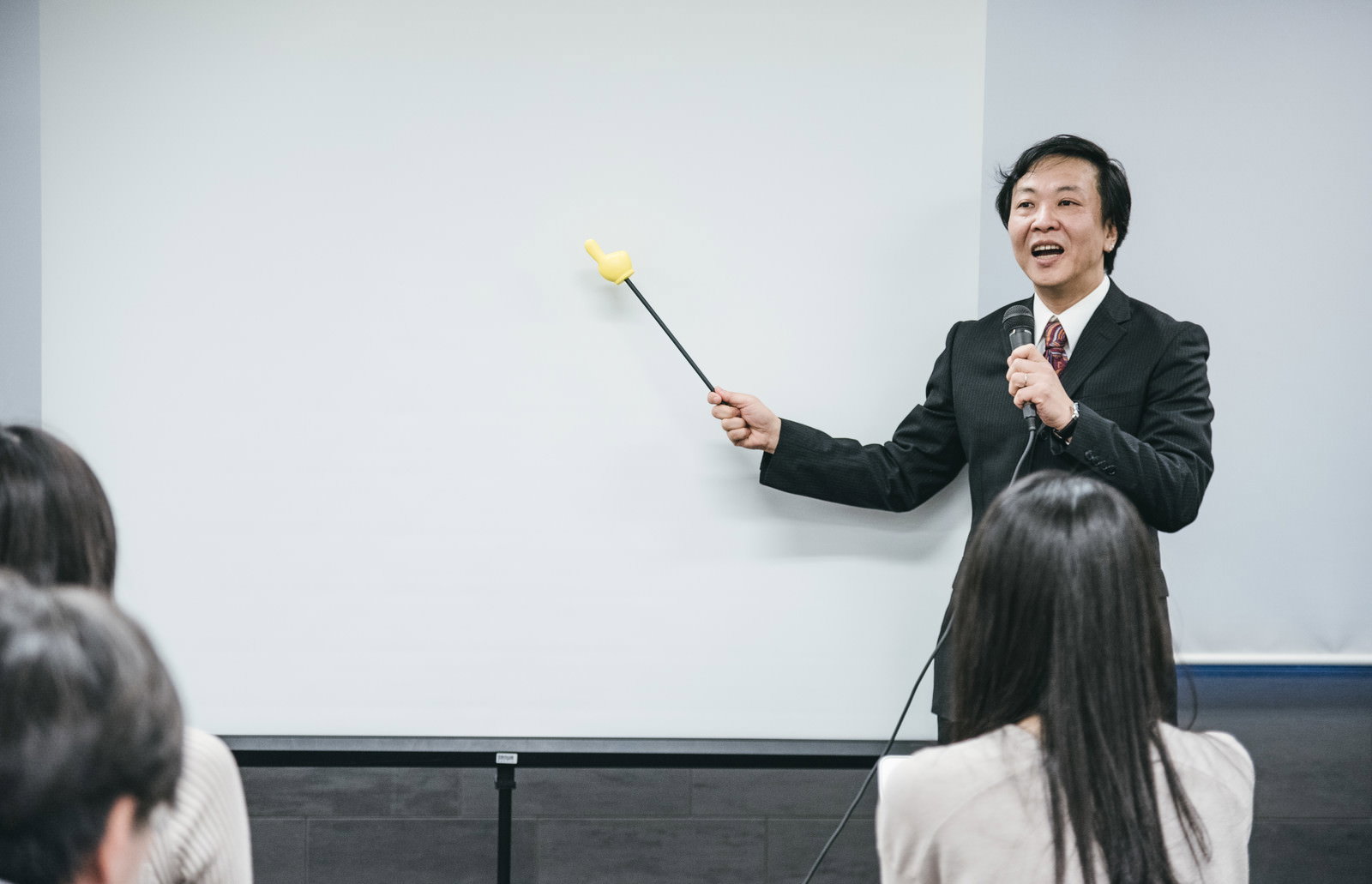 ホワイトボードを見ながら指し棒で説明をするスーツ姿の男性講師［モデル：のせさん］