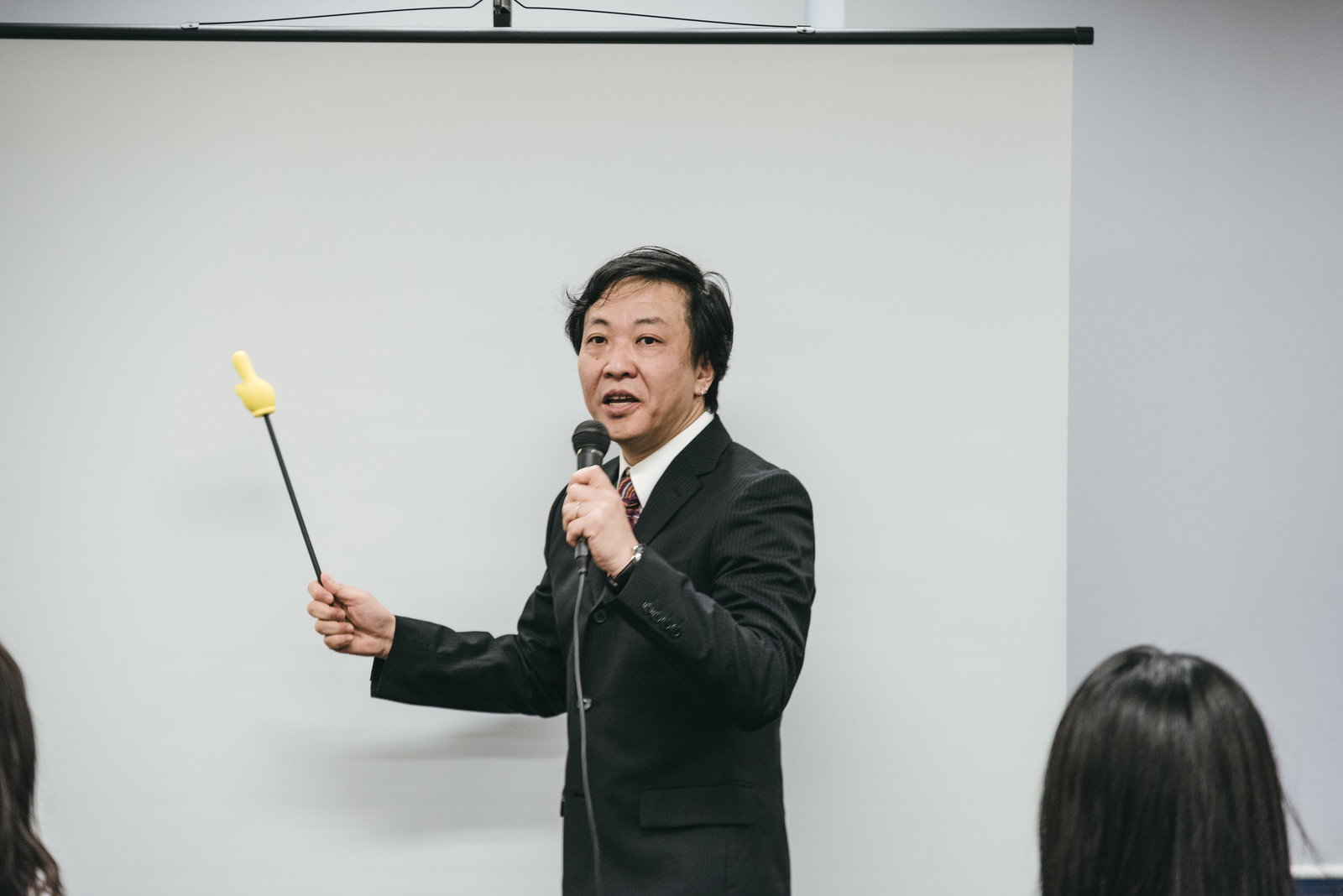 会議室でスーツ姿の上司が指示棒を持ちプレゼンテーションしている［モデル：のせさん］