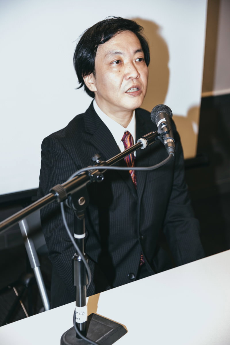 黒いスーツを着た男性が記者会見で複数のマイクの前で謝罪している［モデル：のせさん］