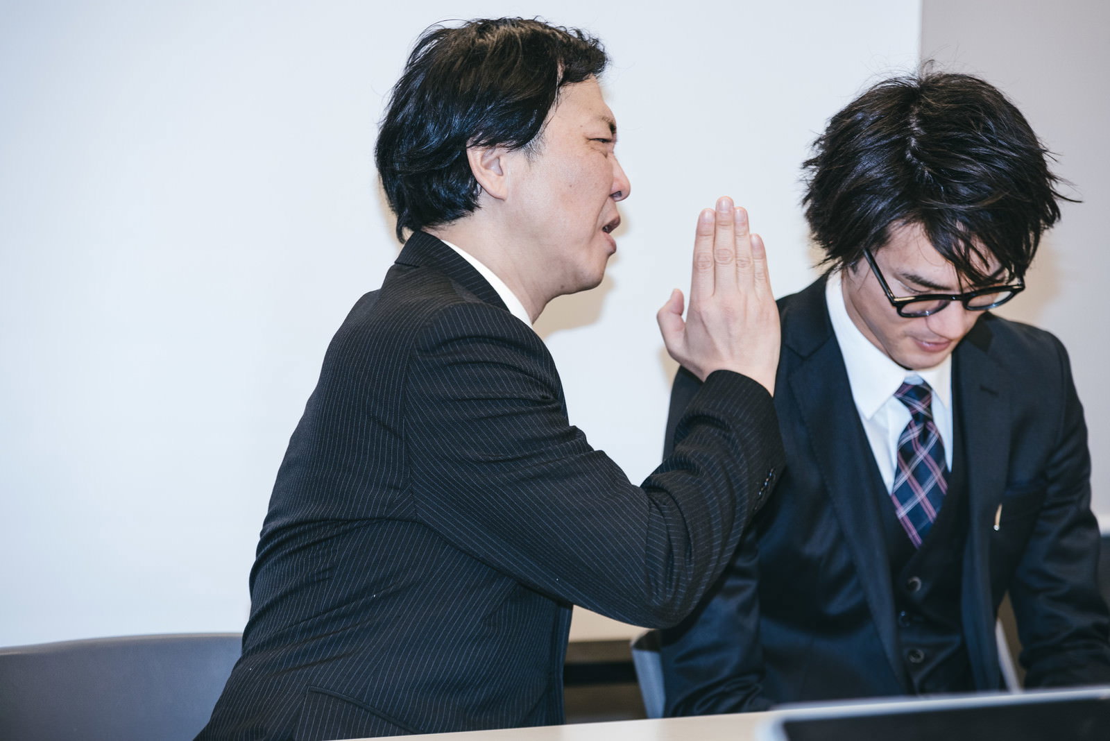 記者会見中にスーツ姿の上司が隣に座る広報担当者に耳打ちしている［モデル：のせさん 村田裕章］