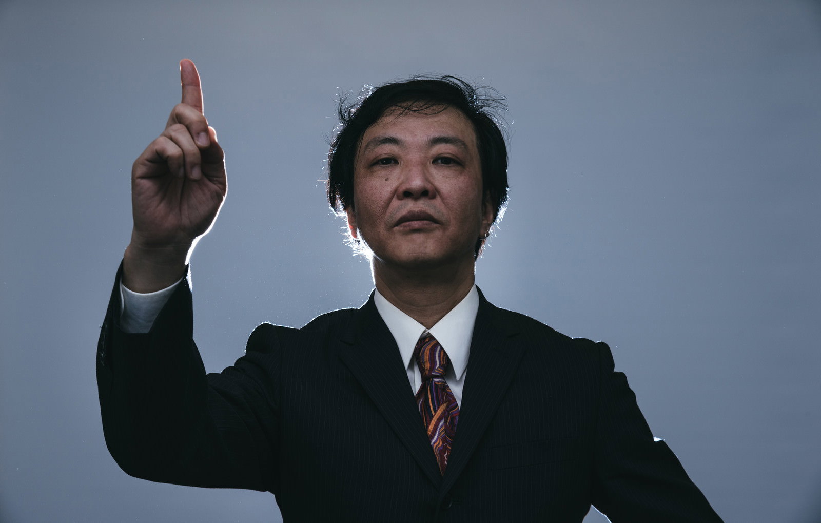 人差し指を立てて上を指差すスーツ姿の男性［モデル：のせさん］