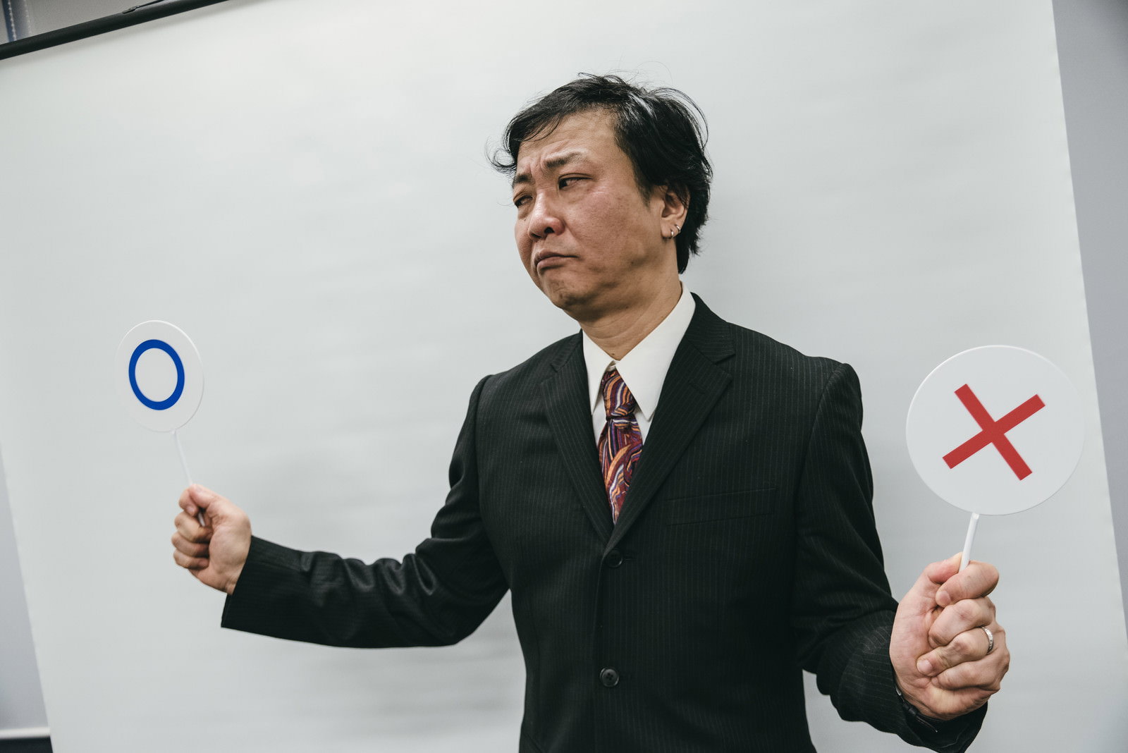○×の札を両手に持ち困った表情を見せるスーツ姿の男性［モデル：のせさん］