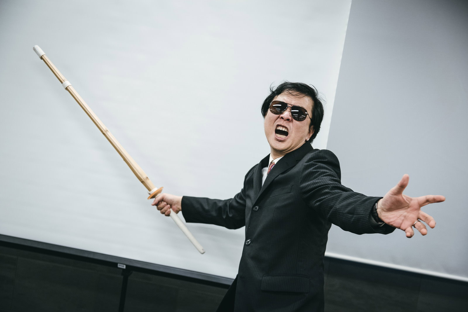 竹刀を持って威圧的なポーズを取るスーツ姿の男性［モデル：のせさん］