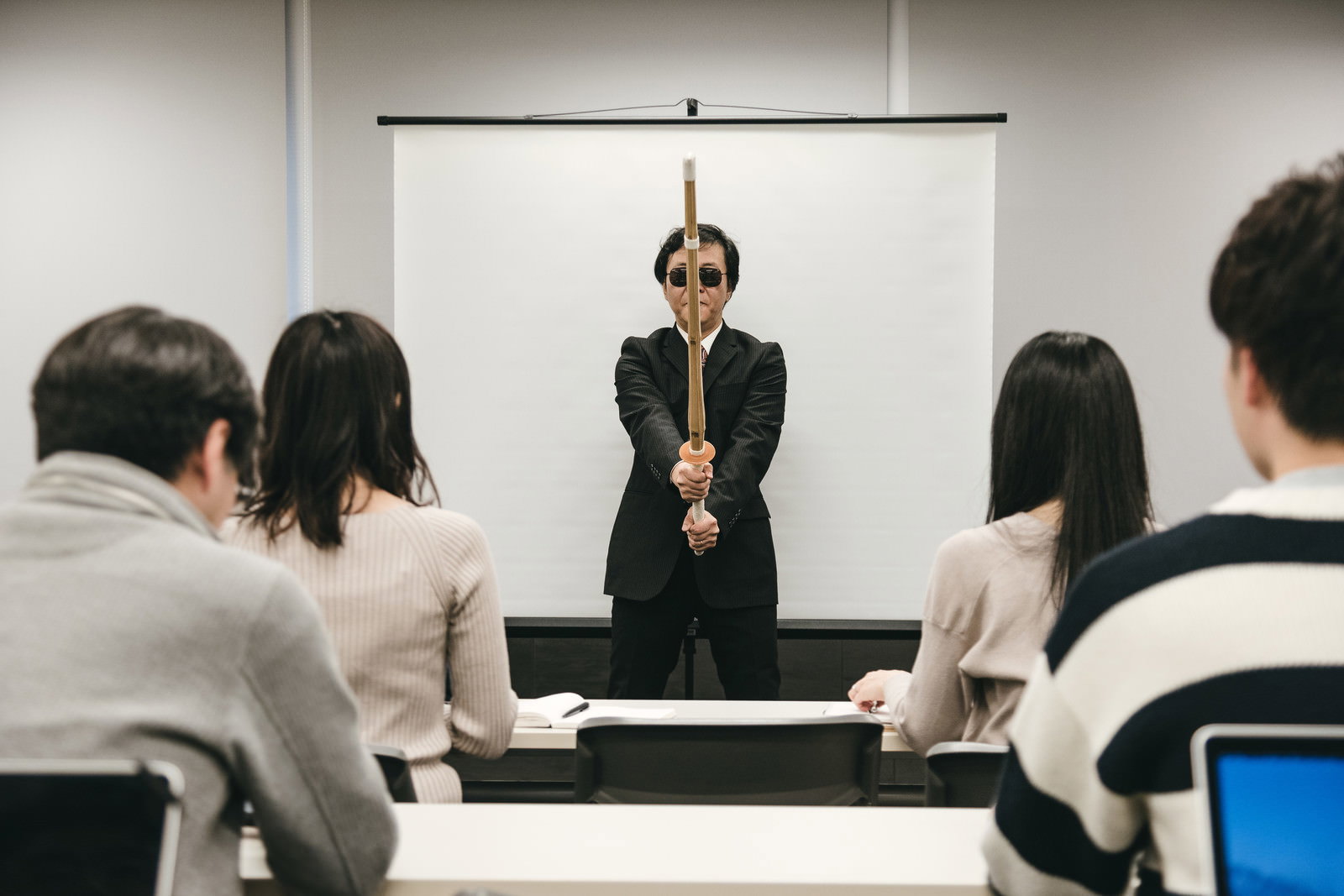 スーツ姿の男性講師が竹刀を持って会議室で指導している［モデル：のせさん］