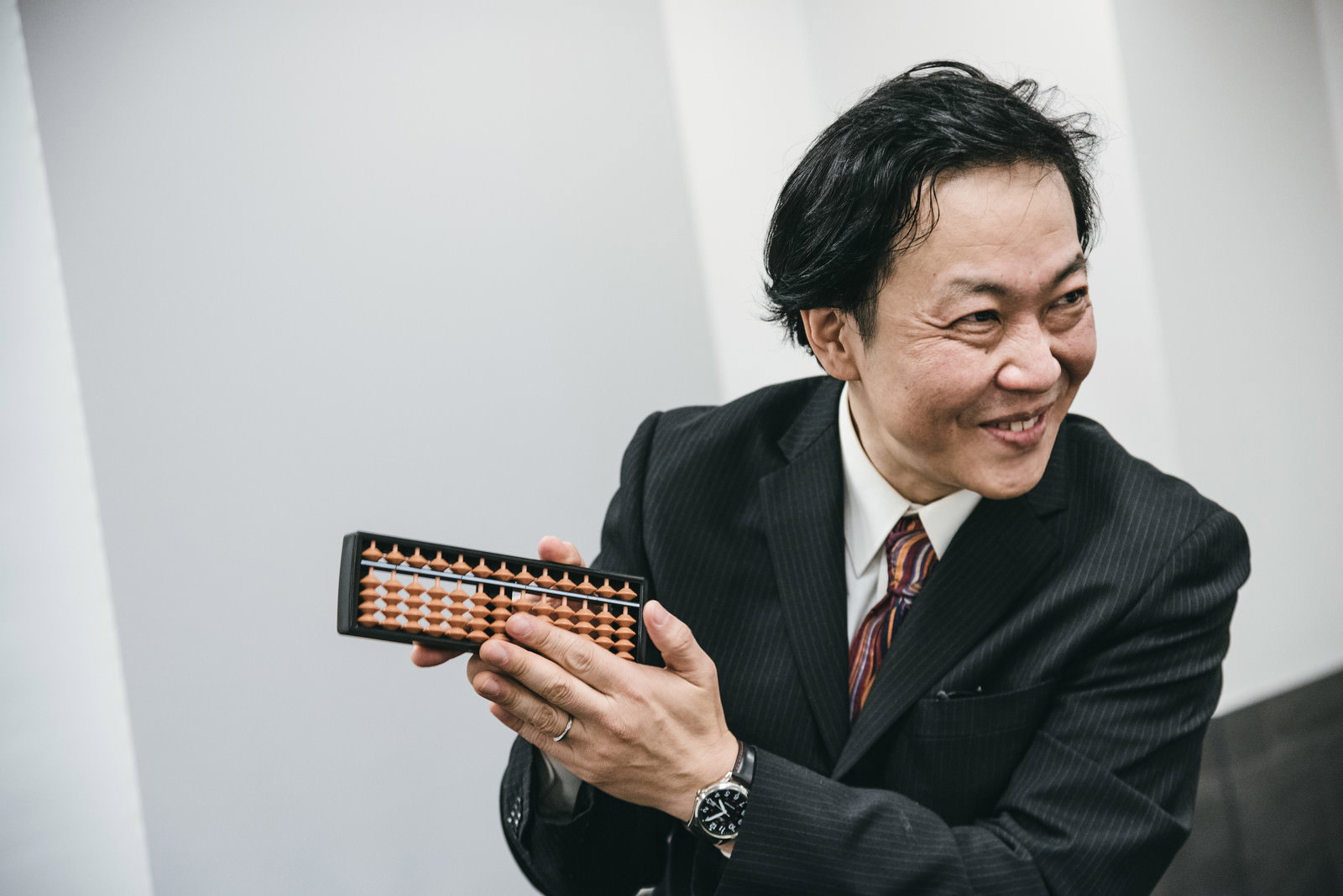 そろばんを持って笑顔を見せる黒いスーツを着た会社員男性［モデル：のせさん］