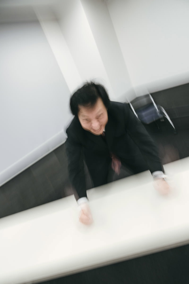 黒いスーツを着た男性が白いテーブルに手をついて怒りの表情を見せている［モデル：のせさん］