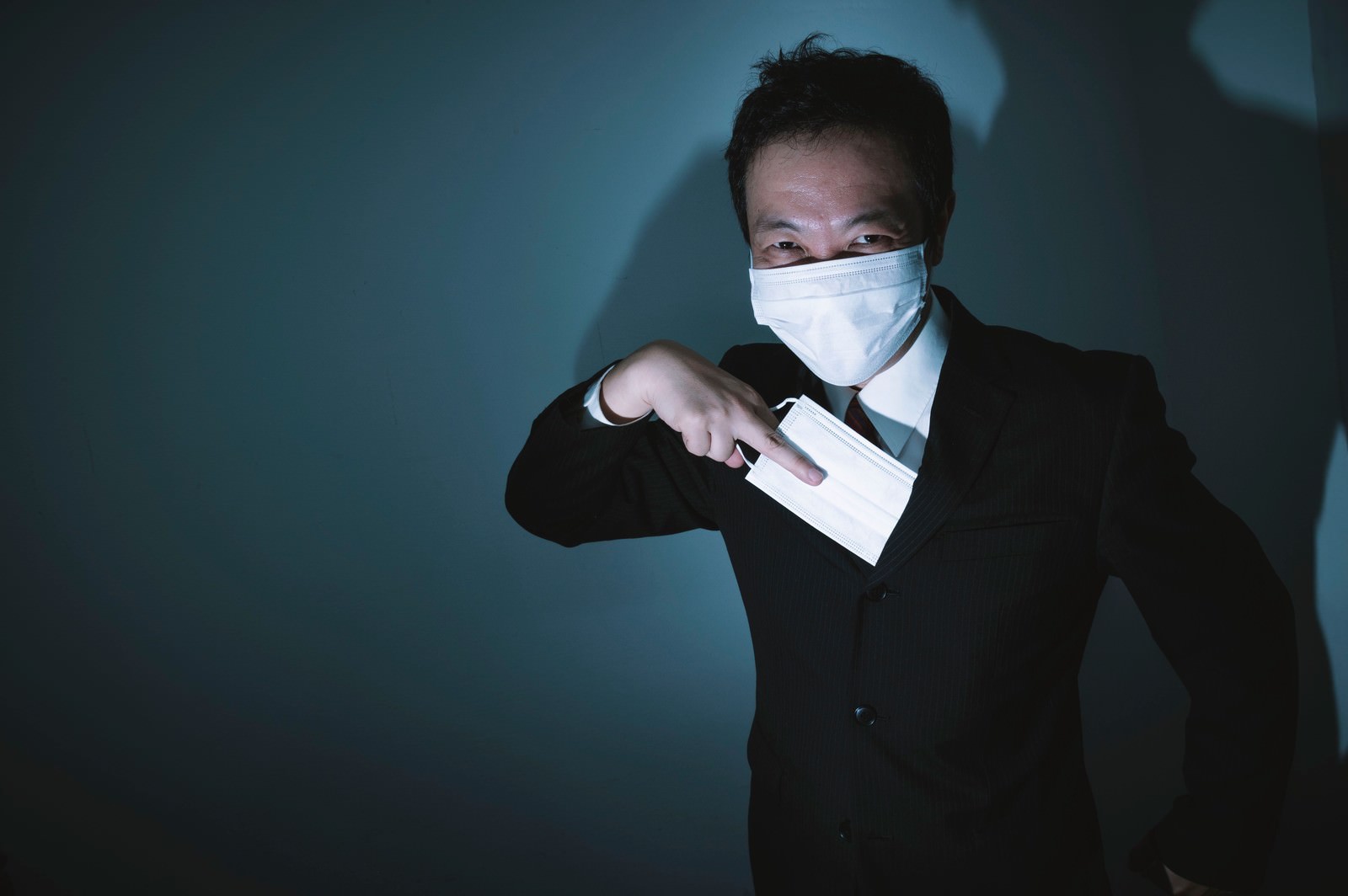 暗い背景でスーツを着た男性がマスクを着用し胸ポケットからマスクを取り出している