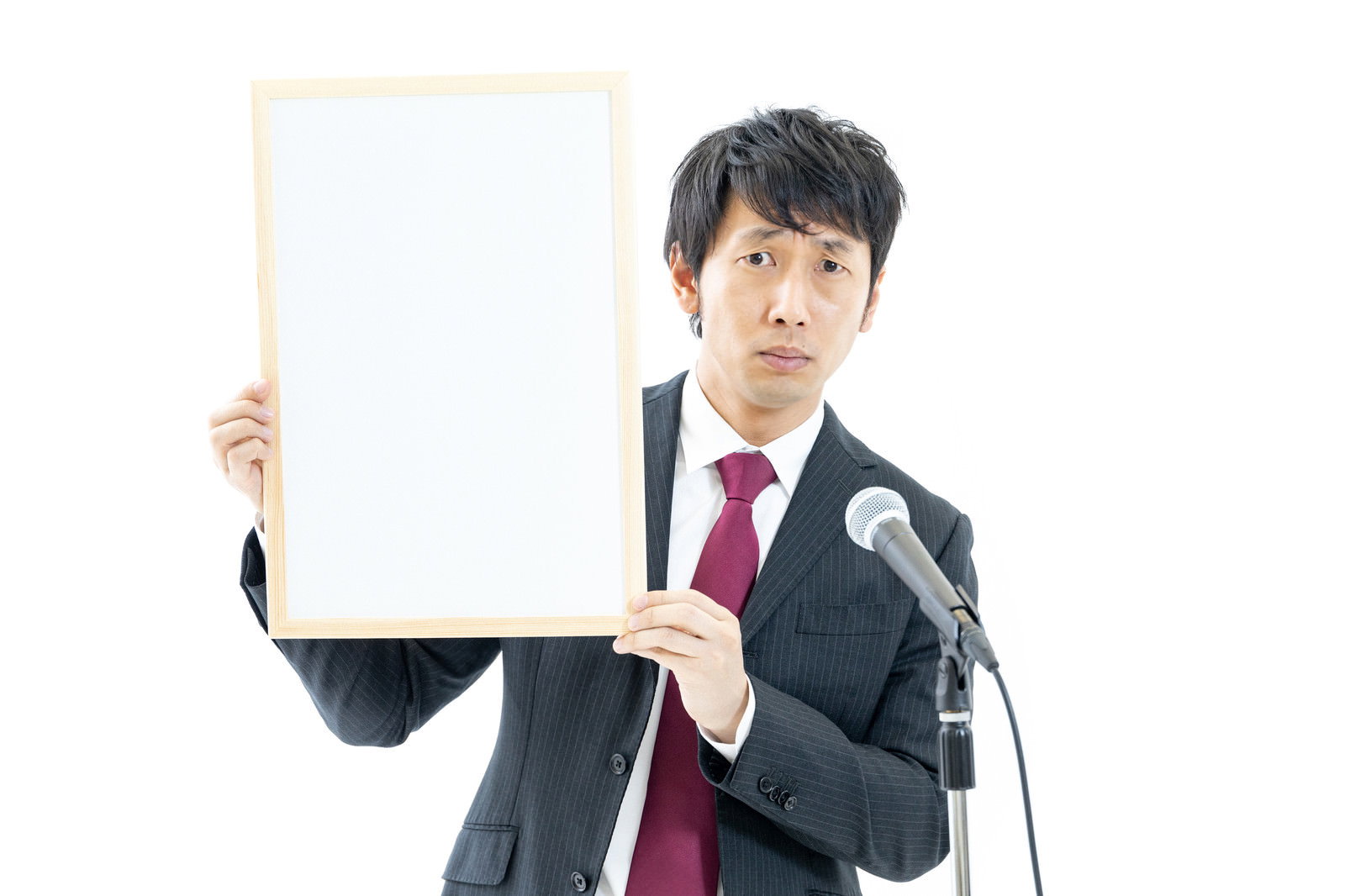 白いボードを持つスーツ姿の男性がマイクの前に立っている［モデル：大川竜弥］