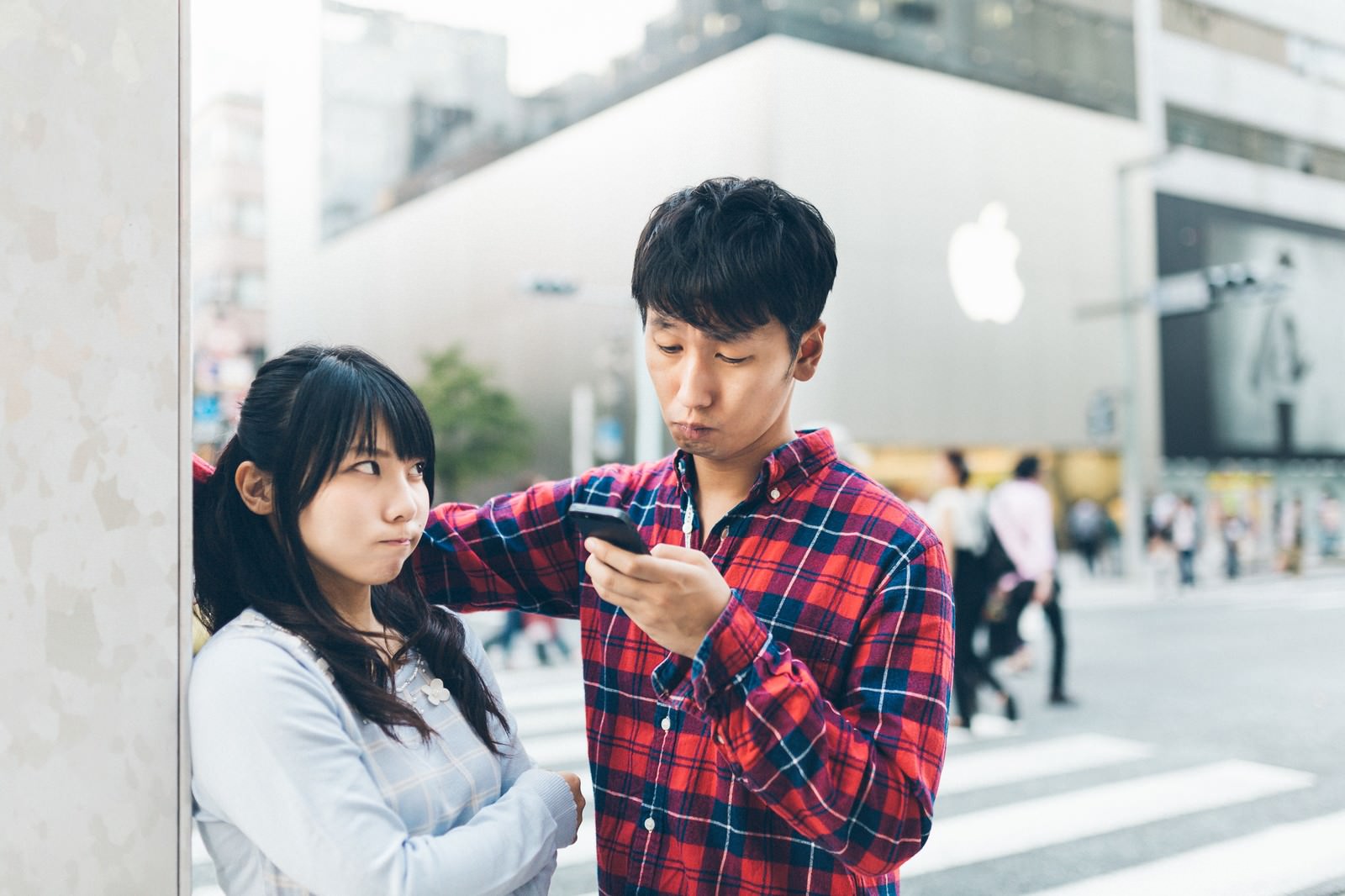 駅前でスマートフォンを見る赤いチェック柄シャツの男性と、腕を組んで怒った表情の白いシャツの女性［モデル：大川竜弥 Lala］