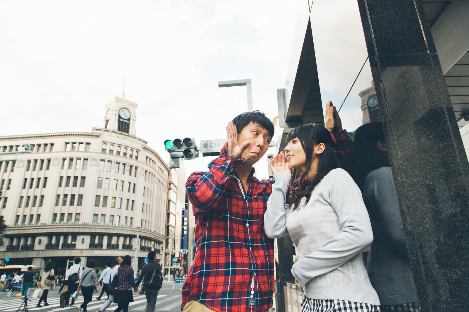 銀座の街中で赤チェックシャツ姿の男性が耳に手をやり、白いトレーナーの女性が笑いながら話しかけている［モデル：大川竜弥 Lala］