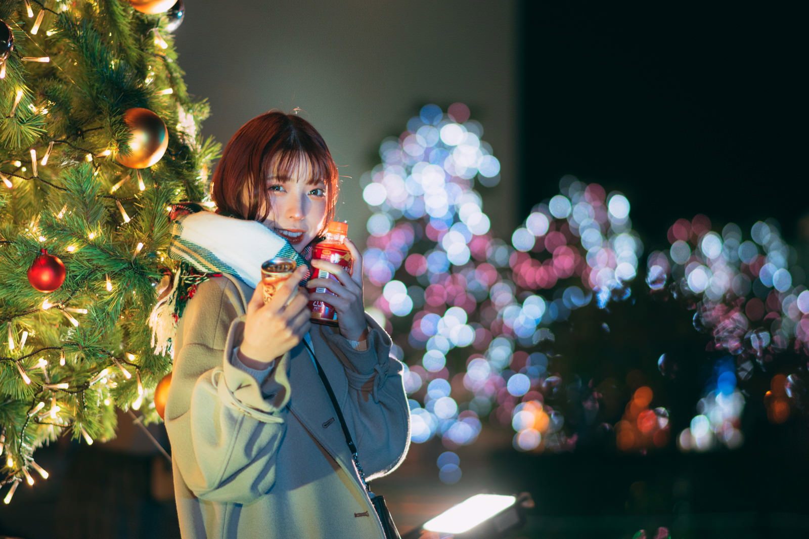 クリスマスツリー前でホットドリンクを持つ女性［モデル：高尾実生］