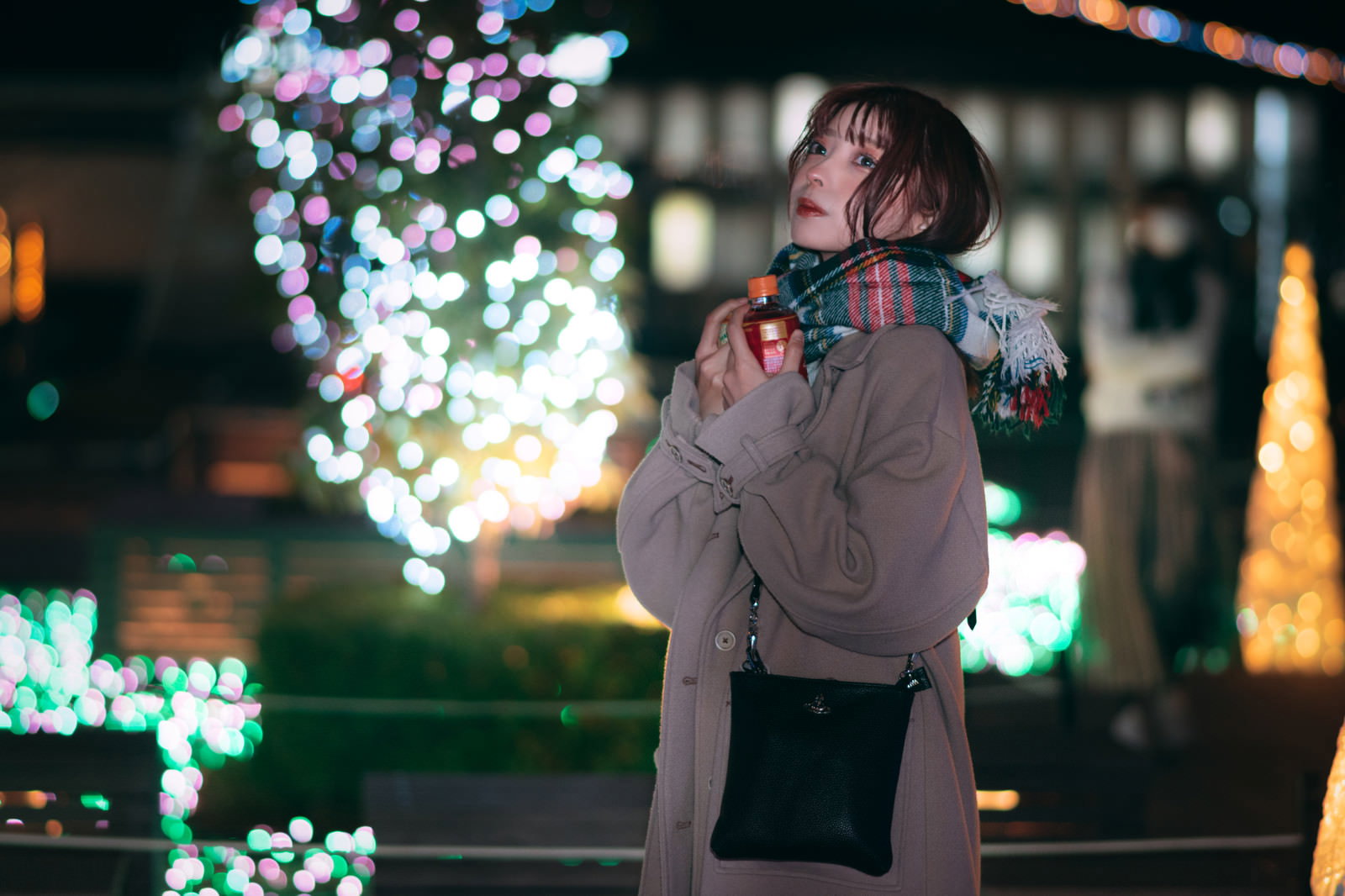 夜のイルミネーションを背景に、マフラーを巻いた女性が立っている［モデル：高尾実生］