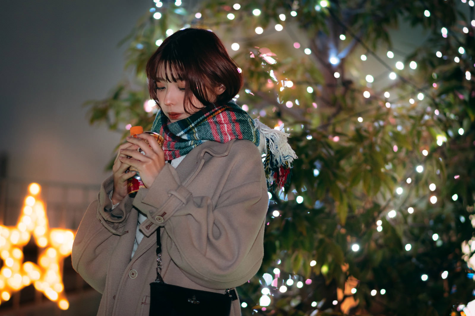 イルミネーションが輝くクリスマスツリーの前で温かい飲み物のカップを持つ女性［モデル：高尾実生］