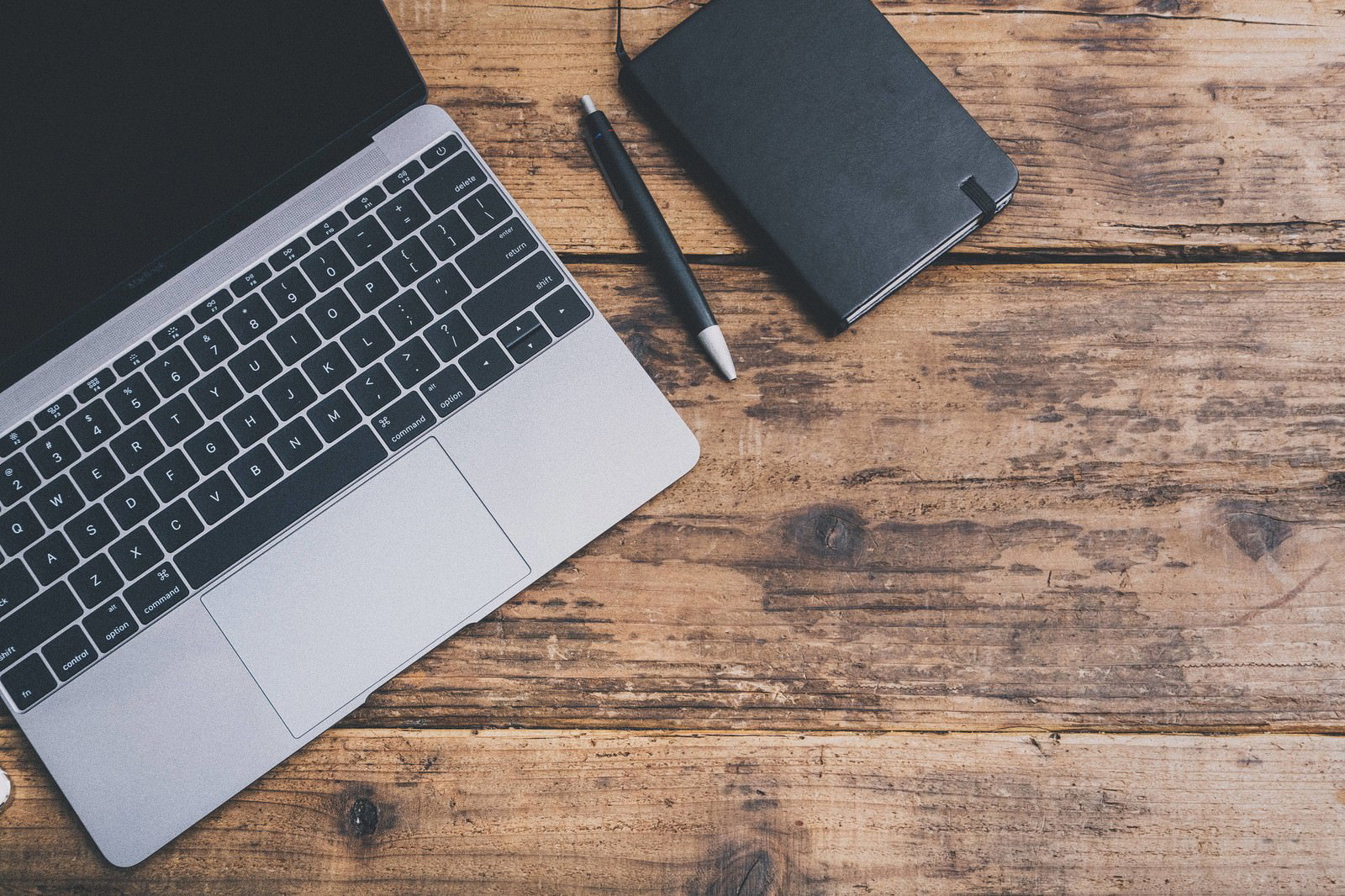 木製デスクの上にMacBookと黒い手帳とペン