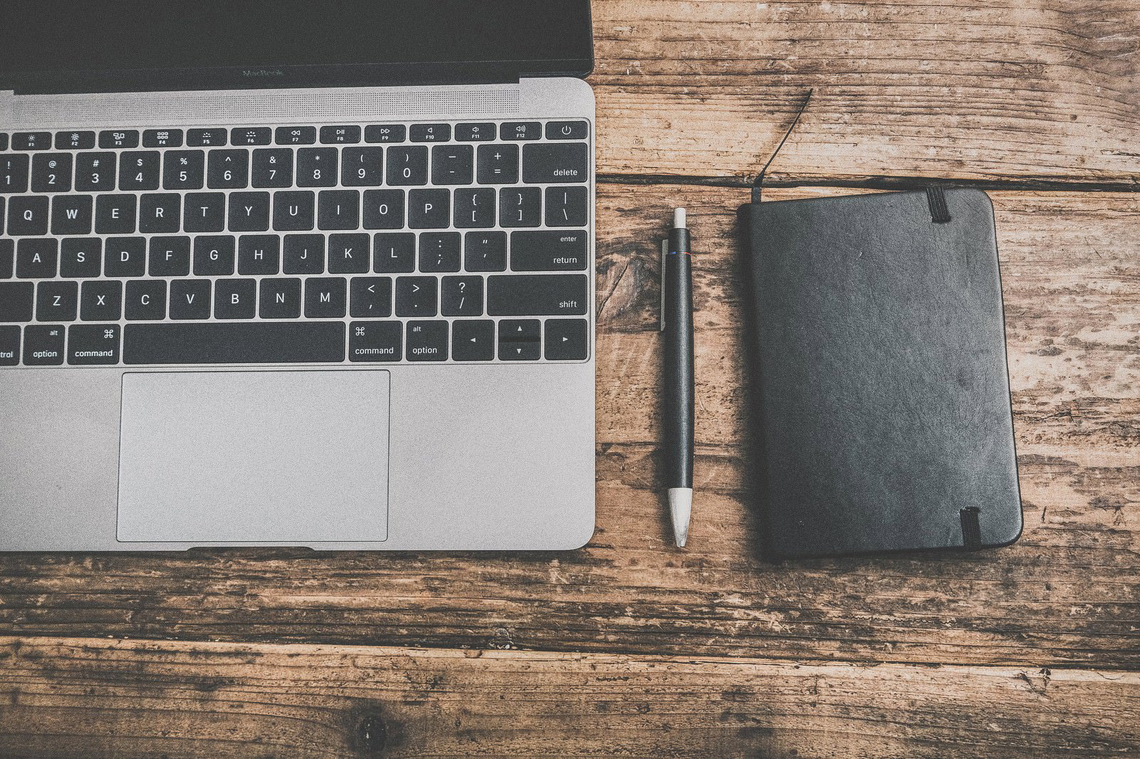 木製テーブルの上のMacBookと黒い手帳とペン