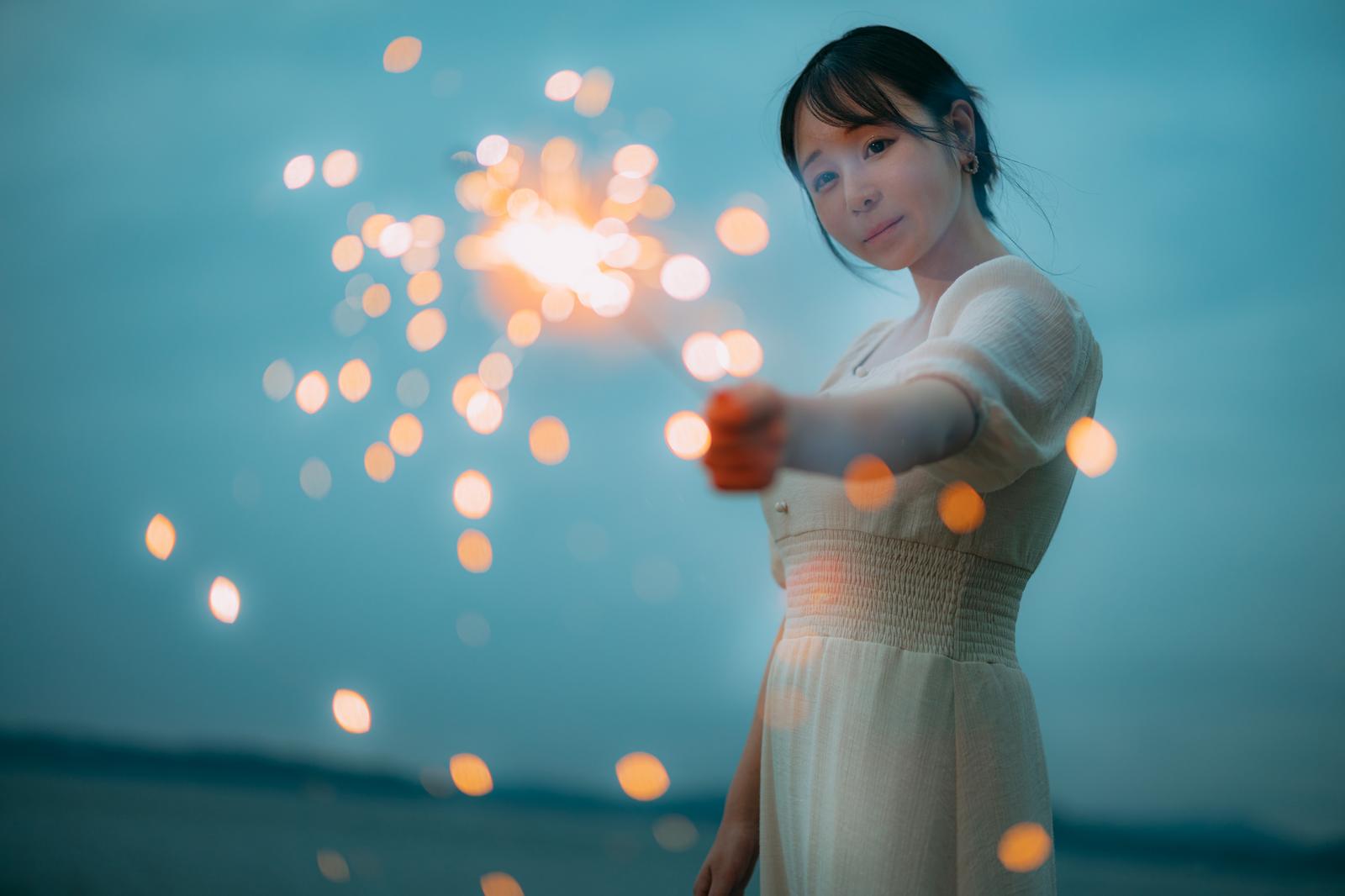 白いワンピースを着た女性が手持ち花火を持ち、夜空に光る火花を見つめている［モデル：茜さや］