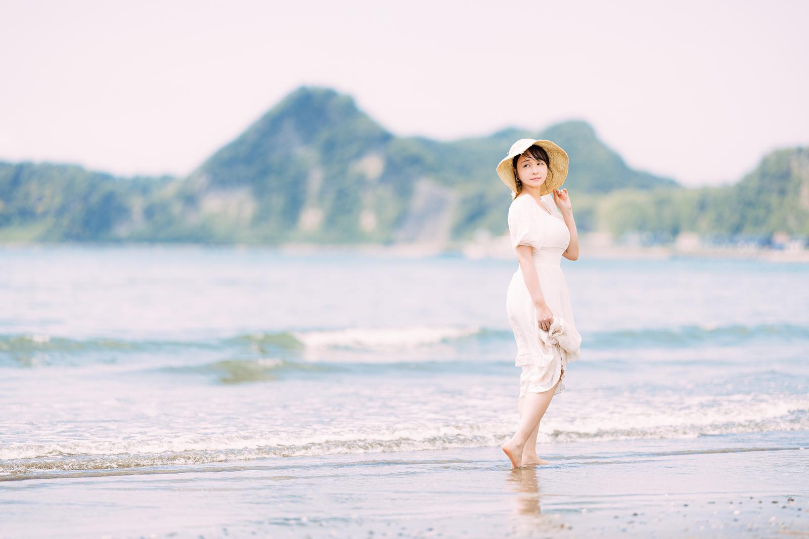 麦わら帽子と白いワンピース姿で波打ち際を歩く女性［モデル：茜さや］