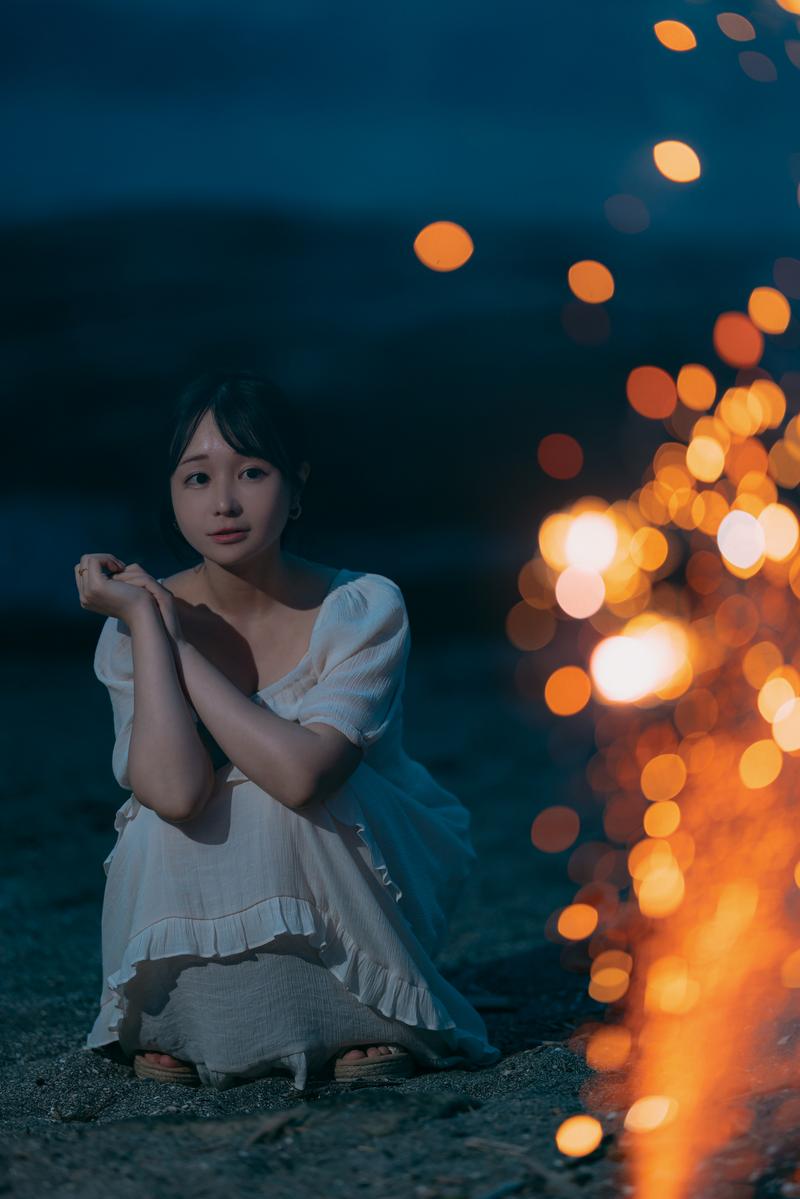 砂浜で光る手持ち花火を持つ女性が夜間に座っている［モデル：茜さや］