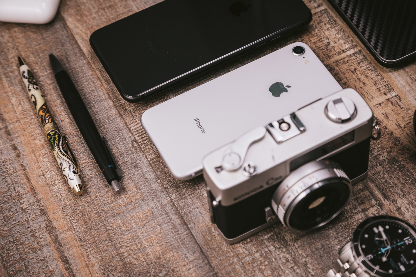 木製テーブルの上にiPhoneとフィルムカメラとペンと腕時計