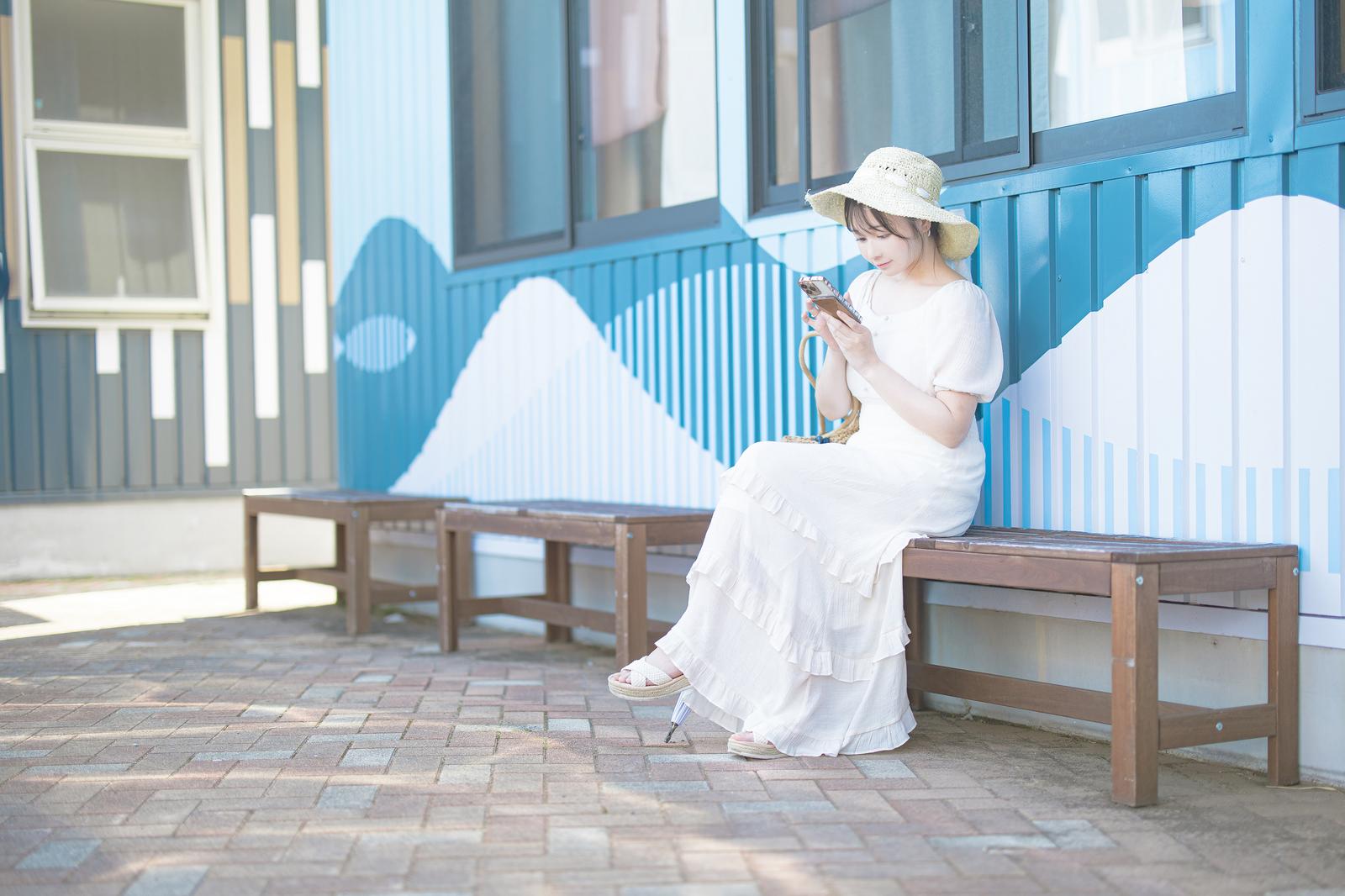 ベンチに座ってスマホを見る麦わら帽子姿の女性［モデル：茜さや］