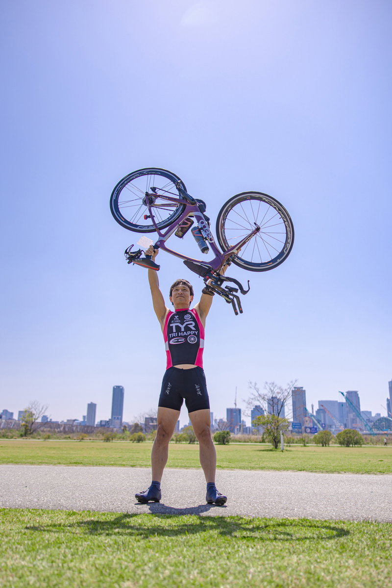 青空の下で自転車を両手で高く掲げて勝利のポーズを決めるトライアスリート［モデル：うえせい］