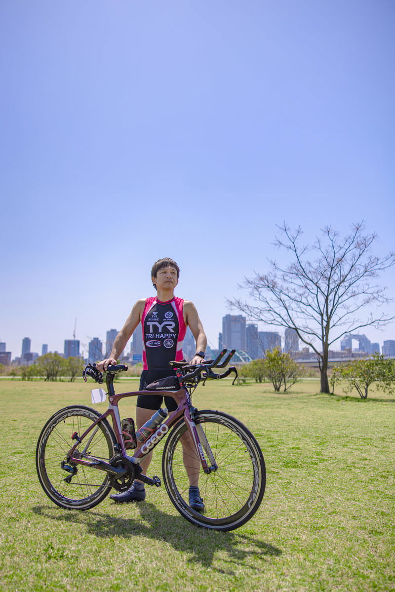 青空の下でロードバイクと一緒に立つトライアスロンウェアの男性選手［モデル：うえせい］