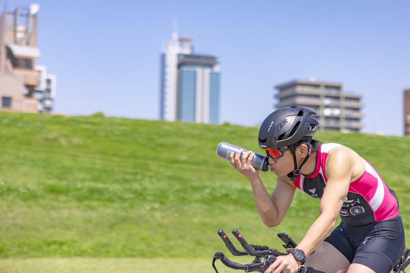 黄色いウェアを着たトライアスリートがバイクに乗りながら水分補給している様子［モデル：うえせい］