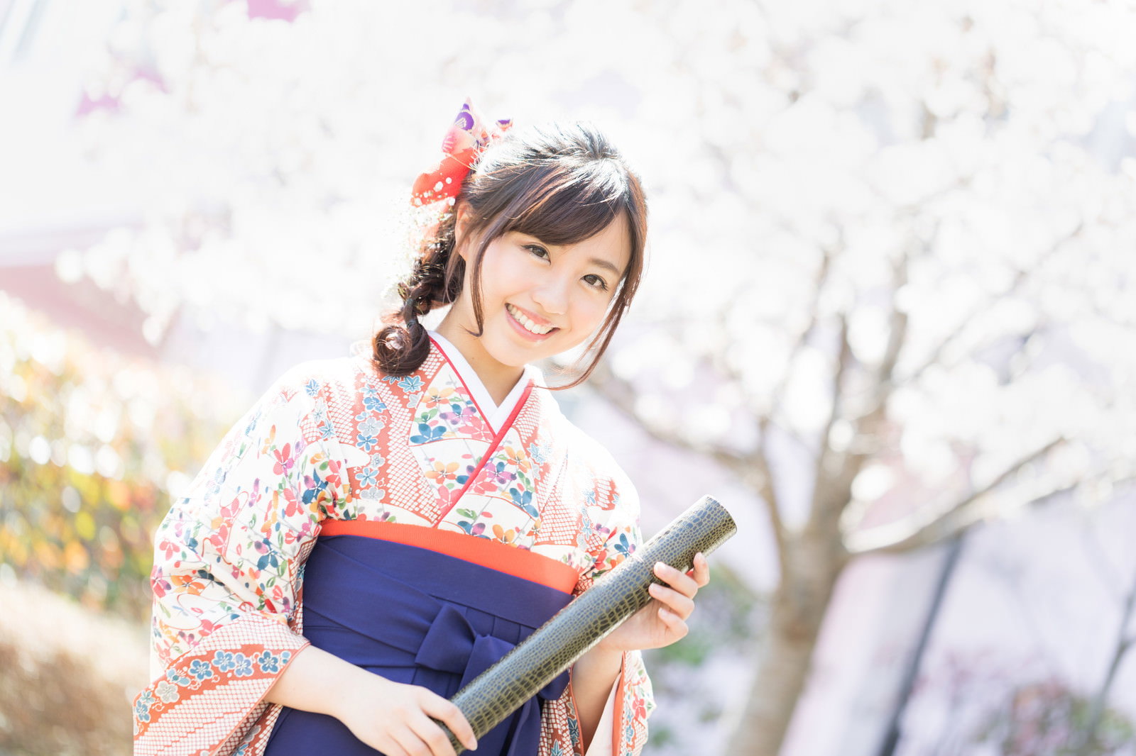 桜の前で卒業証書を持ち微笑む袴姿の女性［モデル：河村友歌］