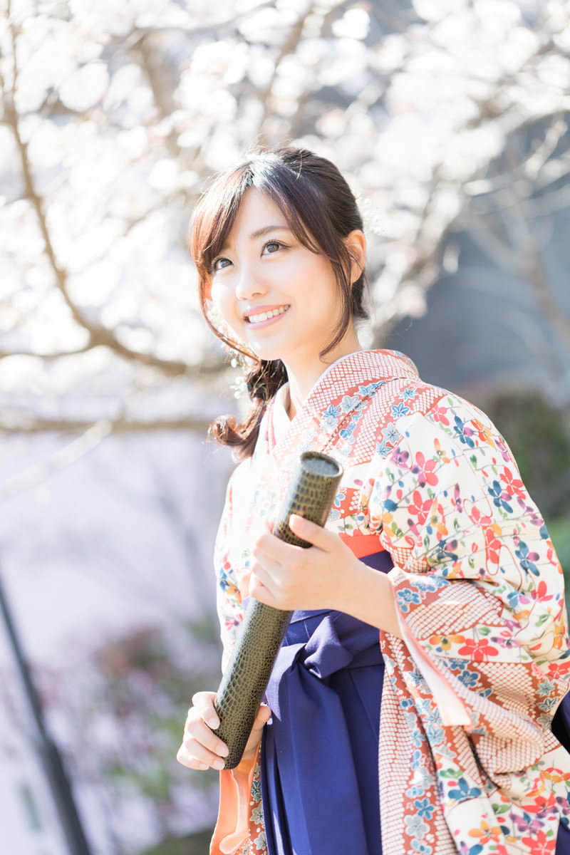 桜の下で卒業証書を持ち微笑む袴姿の女性［モデル：河村友歌］